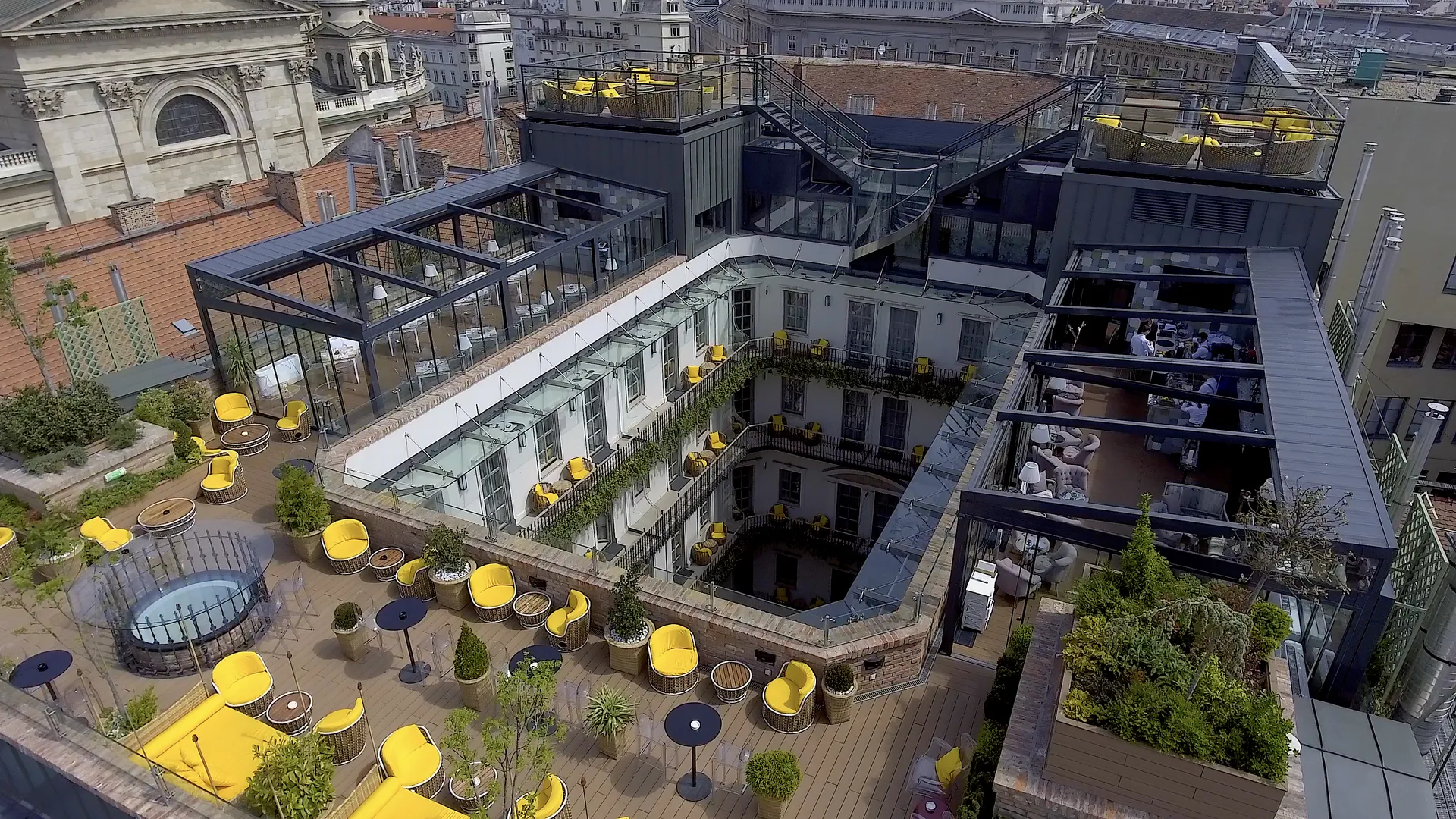 A roof terrace with yellow chairs above a glass-roofed hotel courtyard, city rooftops and facades all around.