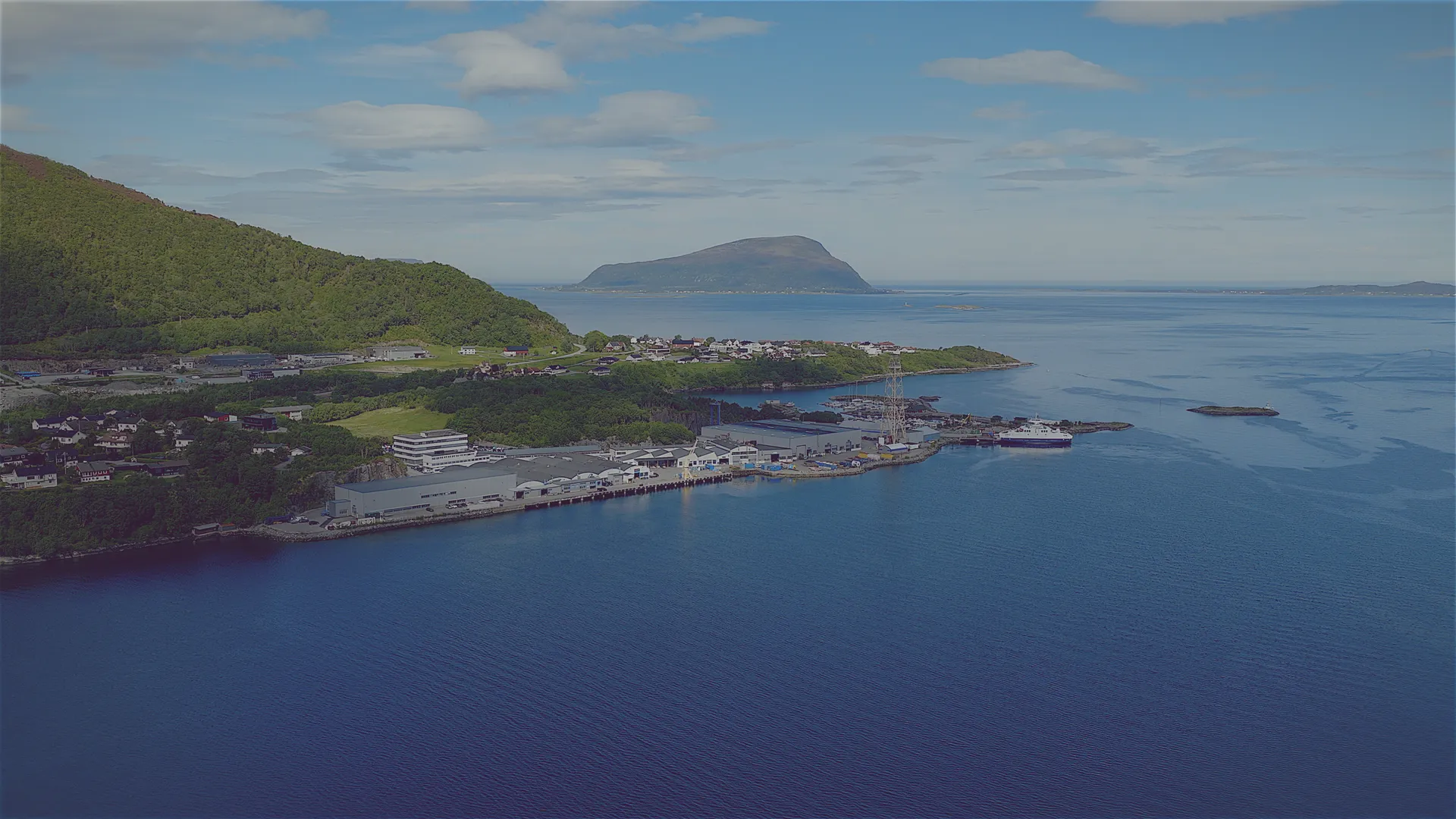 Kystby med industribygninger og havn omgitt av fjord, skogkledde åser og fjell i horisonten.