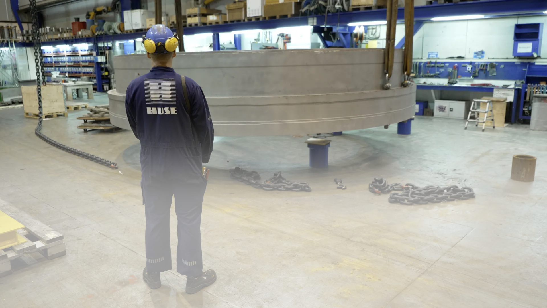 A worker in blue overalls and a hard hat looks at a large steel cylinder suspended in a workshop hall.
