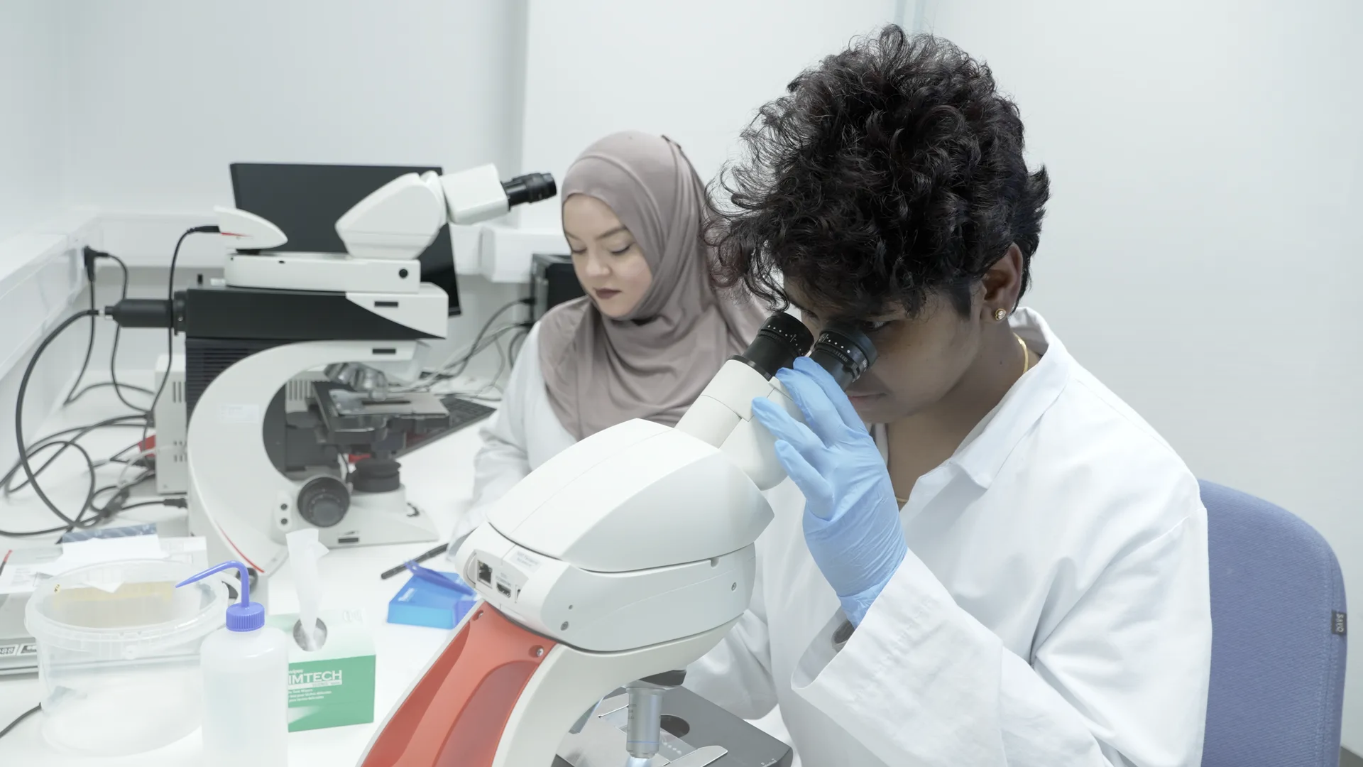 Two people in white coats in a laboratory; the one in front looks into a microscope wearing blue gloves.