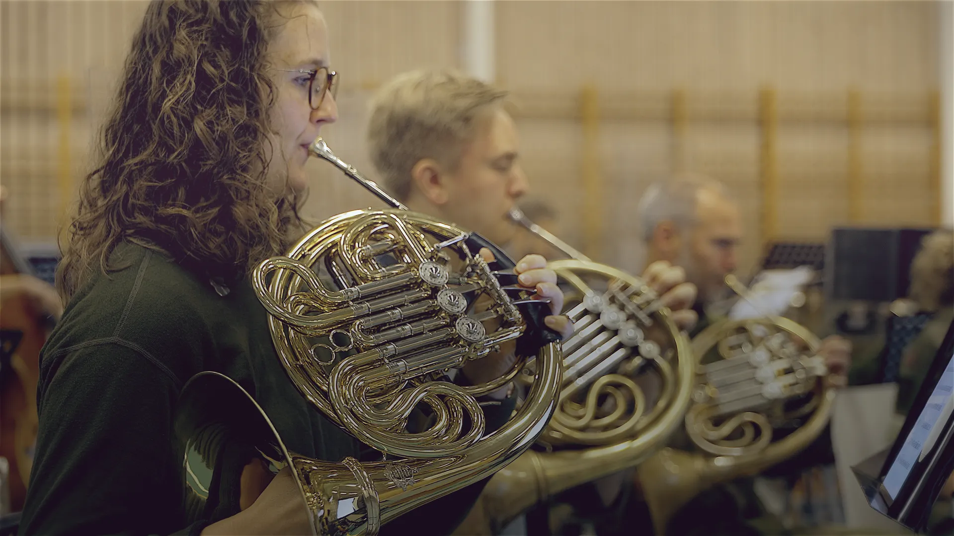 Several French horn players side by side; in front, a woman with curly hair and glasses.