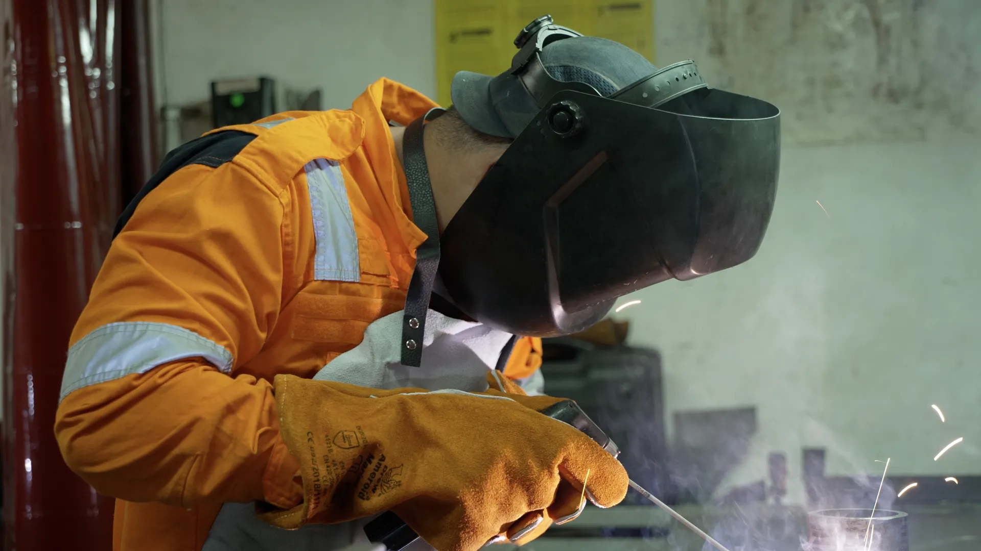 A welder in orange workwear, welding mask and leather gloves bends over the work as sparks fly.