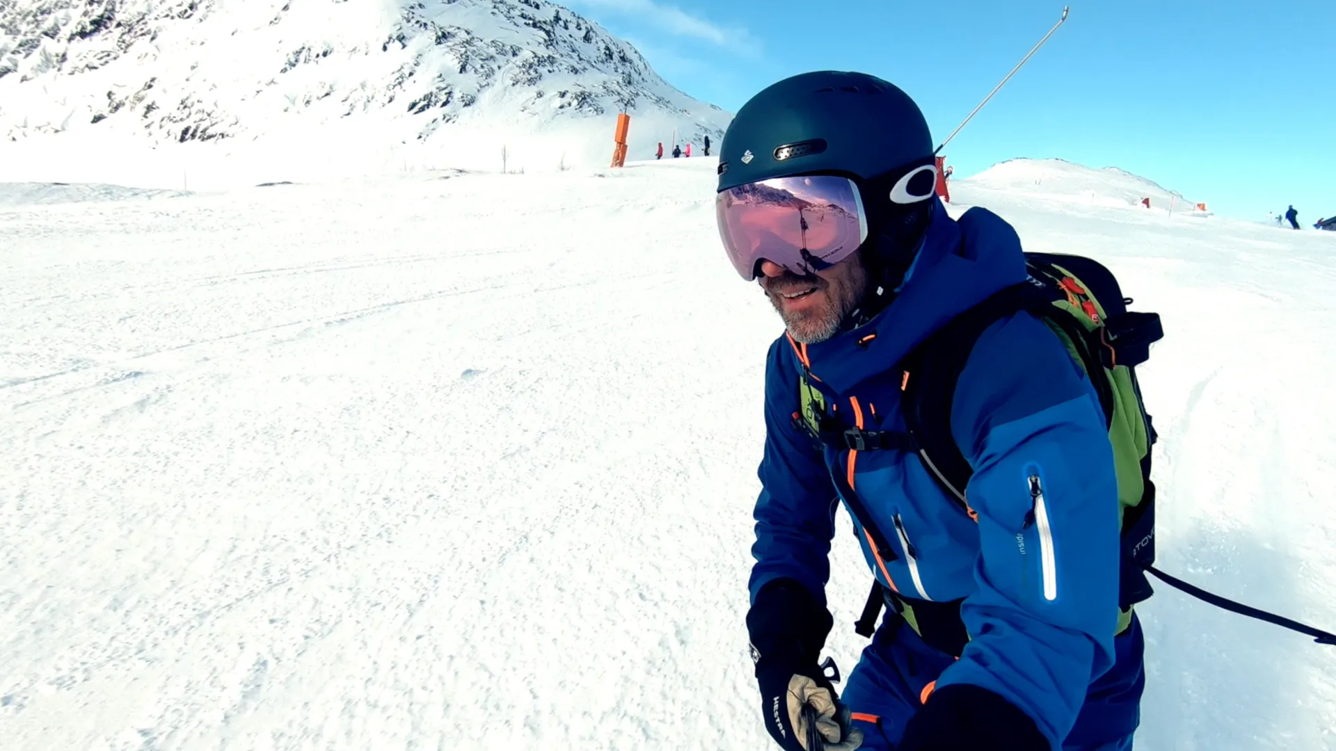 A man in a helmet and ski goggles stands on a groomed piste, snow-covered mountain and blue sky behind.