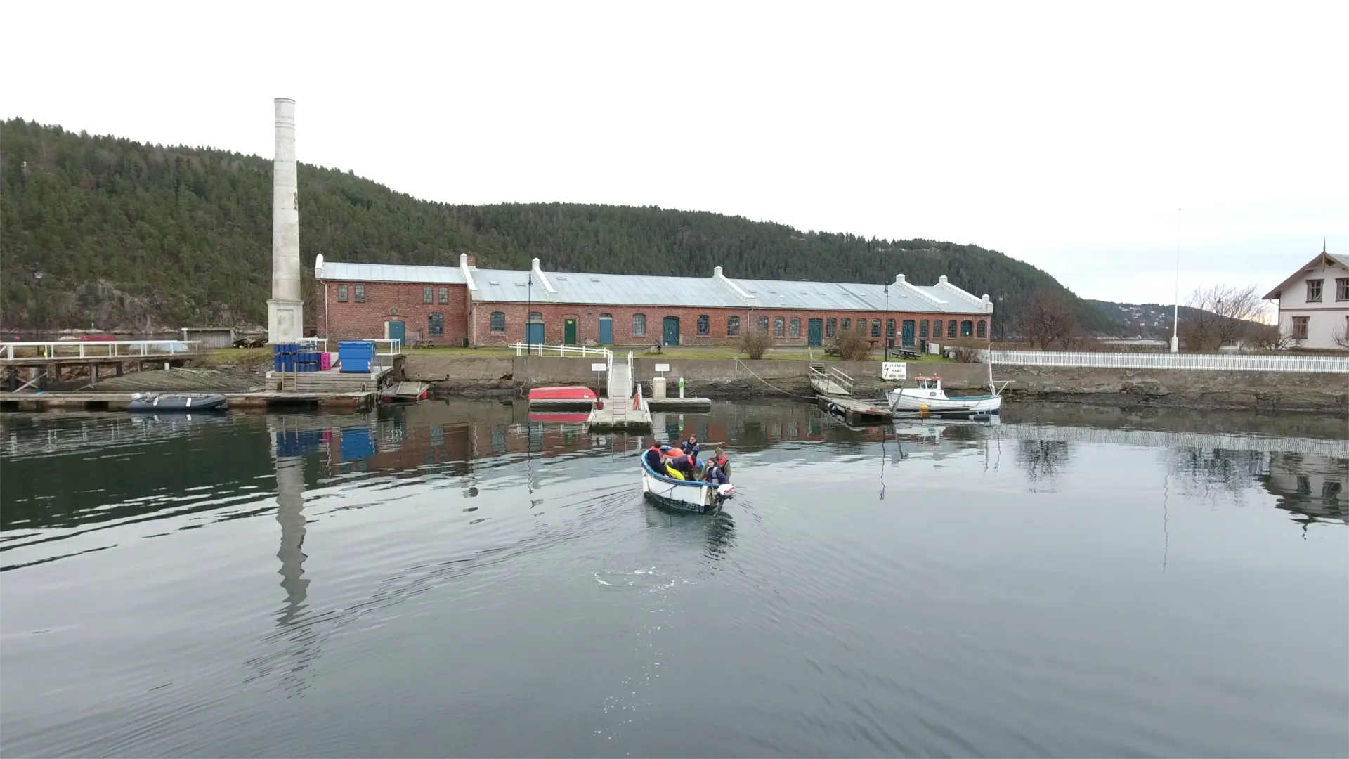 A small boat of people in life jackets crosses calm water toward a long brick building and a tall chimney.